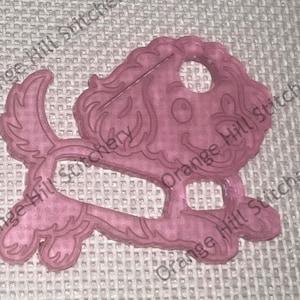 May include: A pink plastic cookie cutter shaped like a cartoon dog with a curly tail and big eyes.