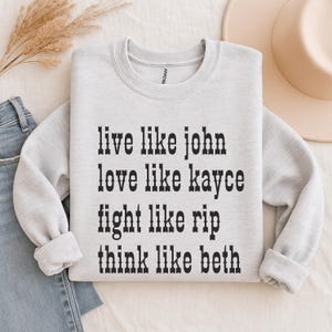 May include: A light gray sweatshirt with black text that reads "live like john, love like kayce, fight like rip, think like beth." The sweatshirt is folded on a white surface, with a pair of blue jeans and a tan hat in the background.