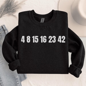 May include: Dark grey sweatshirt featuring the numbers 4 8 15 16 23 42 in white. The garment is folded, with the sleeves neatly tucked in. The Gildan brand label is visible on the inside of the collar.
