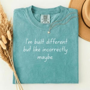 Könnte beinhalten: Ein türkisfarbenes Comfort Colors T-Shirt mit dem Text "I'm built different but like incorrectly maybe." Eine silberne Halskette mit Sternanhänger liegt auf dem Shirt. Ein brauner Hut ist im Hintergrund zu sehen.
