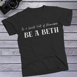 May include: Black t-shirt with the text "In a World Full of Princesses BE A BETH" in white. The shirt is made of a soft material and is displayed on a wooden surface. A camera is visible in the top left corner.