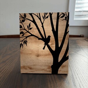Pu&ograve; includere: Un'opera d'arte murale in legno con una silhouette nera di un albero e un uccello. L'albero ha rami con foglie e l'uccello &egrave; appollaiato su un ramo.