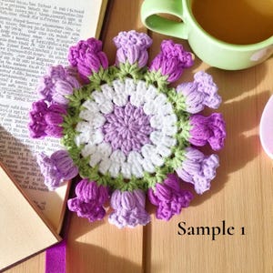May include: A handmade crochet coaster with a floral design. The coaster features a central circle of white and lavender, surrounded by green leaves and purple flower-shaped edges. The text "Sample 1" is visible.