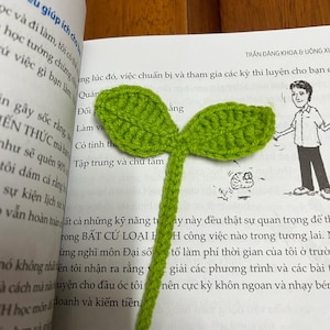 Op de afbeelding: Een handgemaakte groene gehaakte bladwijzer in de vorm van een spruit, met twee bladeren en een stengel. De bladwijzer ligt op een open boek met Vietnamese tekst.
