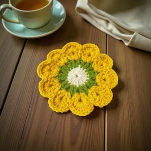 May include: A handmade, yellow crochet flower coaster with green and white accents. The coaster is placed on a wooden surface, with a cup of tea and a saucer in the background. A folded beige cloth is also visible.