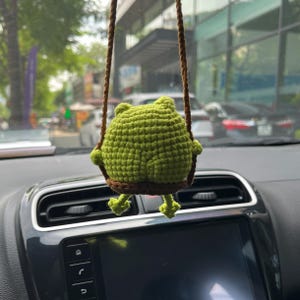 May include: A green crocheted frog ornament hanging from brown twine. The frog is sitting on a brown swing, with its legs dangling. The ornament is likely for a car or home decor.
