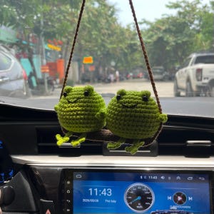 May include: Two round, bright green crocheted frogs sit side-by-side on a small, brown, horizontal branch. The frogs have small black bead eyes and are suspended by a thin brown cord, designed to hang from a vehicle's rearview mirror.