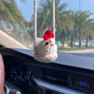 May include: A handmade crochet chicken car charm. The chicken is white with a red comb and a light orange beak. It hangs from a beige string. The charm is hanging in front of a car dashboard.