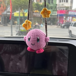 May include: A handmade crochet car charm featuring a pink character with blue eyes and a smiling face. The charm is suspended by brown yarn and adorned with yellow star-shaped accents. The charm is hanging in front of a car dashboard.