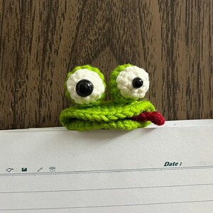 Op de afbeelding: Een handgemaakte groene gehaakte kikker bladwijzer met witte en zwarte ogen en een rode tong. De bladwijzer is geplaatst op gelijnd papier met het woord "Date:" erop gedrukt. De achtergrond is een houten oppervlak.