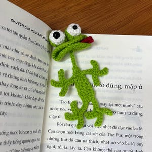 May include: A handmade green crochet bookmark shaped like a lizard, with white and black eyes and a red tongue. The bookmark is placed in an open book with Vietnamese text.
