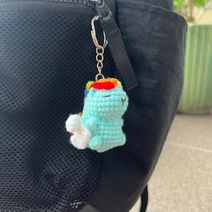 May include: A handmade crochet keychain featuring a light blue dinosaur with a rainbow-colored headpiece and a white cloud-like detail. The keychain is attached to a silver chain and a black backpack.