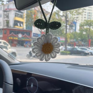 May include: A handmade crochet daisy car charm. The daisy has white petals and a yellow center. Two green leaves and a green string are attached to the daisy. The charm is hanging from a car's rearview mirror.