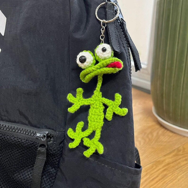 Op de afbeelding: Een handgemaakte groene gehaakte kikker sleutelhanger met witte ogen en een rode mond. De kikker is bevestigd aan een zilveren sleutelring en hangt aan een zwarte rugzak. De armen en benen van de kikker zijn uitgestrekt.