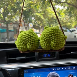 Peut inclure: Deux figurines crochetées vert citron en forme de fesses arrondies sont fixées sur une balançoire en crochet marron. La balançoire est suspendue par deux cordons en laine marron, conçus pour être accrochés au rétroviseur d'un véhicule.