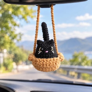 Può includere: Un ornamento a forma di gatto nero all'uncinetto con occhi grandi, naso rosa e baffi bianchi, seduto all'interno di un cestino color marrone chiaro all'uncinetto. Il cestino è sospeso da due cinghie, progettato per lo specchietto retrovisore.