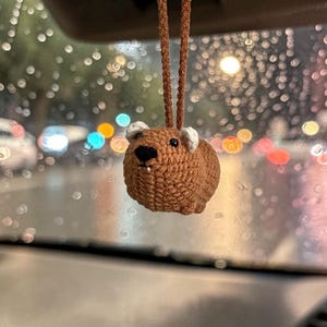 Op de afbeelding: Een klein, bruin, gehaakt capybara-ornament dat aan een bruin koord hangt. De capybara heeft een rond lichaam, kleine witte oren, zwarte kraaloogjes, een zwarte neus en twee piepkleine witte tandjes zichtbaar aan de voorzijde van de mond.