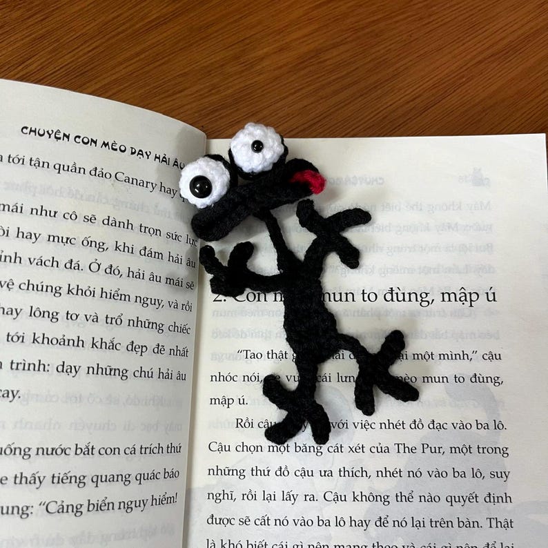 Op de afbeelding: Handgemaakte zwarte gehaakte bladwijzer in de vorm van een grillig wezen met grote witte ogen en een rode mond. De bladwijzer ligt op een open boek met Vietnamese tekst.