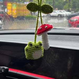May include: A handmade crochet car charm featuring a green frog, a red and white mushroom, and green leaves. The frog has a white belly and black eyes. The mushroom has white spots. The charm is suspended by a green string.