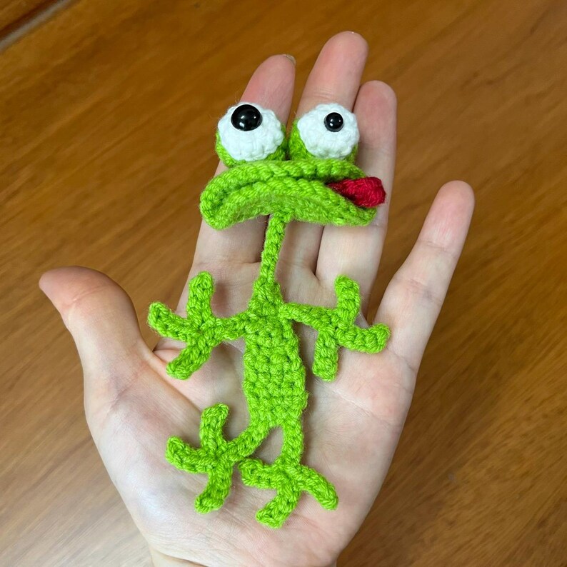 Op de afbeelding: Een handgemaakte groene gehaakte kikker met witte ogen en een rode tong. De kikker heeft uitgestrekte armen en benen en wordt in een hand gehouden. De kikker is een grillig, handgemaakt item.