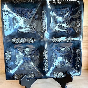 Puede incluir: Un plato de servir de cerámica azul con cuatro secciones divididas. Cada sección tiene un diseño en relieve de hojas y bayas alrededor del borde. El plato está sobre un soporte negro.