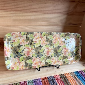 Könnte beinhalten: Rechteckige Serviertablett mit floralem Muster. Das Tablett hat eine glänzende Oberfläche und ist mit rosa und weißen Blumen, grünen Blättern und goldenen Akzenten auf dunklem Hintergrund verziert. Das Tablett wird auf einem schwarzen Metallständer präsentiert.