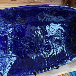 Könnte beinhalten: Ein rechteckiger, tiefblauer Keramik-Servierteller mit einem strukturierten, organischen Muster. Die glänzende Oberfläche reflektiert das Licht und hebt die komplizierten Details des Designs hervor. Der Teller wird von einem schwarzen Metallständer getragen, der seine dekorativen und funktionalen Eigenschaften zeigt.