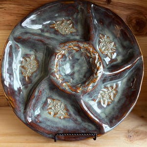 May include: A handcrafted ceramic serving platter with a divided design. The platter features a glossy, mottled glaze in shades of blue, brown, and green. Decorative floral relief patterns are placed around the edges and center. The platter is supported by a black metal stand.