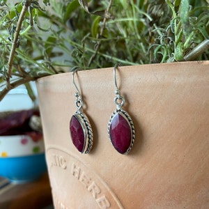Könnte beinhalten: Ein Paar silberne Ohrhänger mit einem dunkelroten Edelstein in Ovalform. Die Ohrringe haben ein geflochtenes Design um den Edelstein herum.