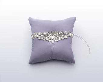 May include: A lavender ring pillow with a decorative silver and crystal band. The band features a central heart-shaped crystal and leaf-shaped accents. Silver cords hang from the band. The pillow is square and sits on a white background.