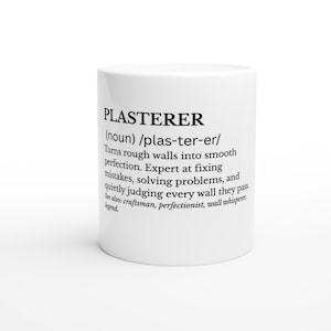 May include: A white ceramic mug featuring black printed text that defines the word PLASTERER as a noun, describing someone who turns rough walls into smooth perfection, fixes mistakes, solves problems, and judges walls, with additional see also terms.