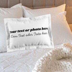 Könnte beinhalten: Weißes Kissen mit dem Text "Your text or photo here Dein Text oder Foto hier" auf einem Bett mit weißer Bettwäsche und einem cremefarbenen Strickwurf. Mehrere weiße Kissen sind hinter dem vorgestellten Kissen angeordnet.