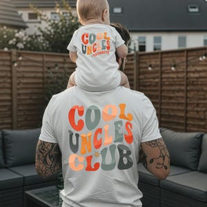 Könnte beinhalten: Weiße T-Shirts mit dem Text "COOL UNCLES CLUB" und "COOL UNCLES FAVORITE" in Retro-Schrift in Orange, Türkis und Gelb. Ein Baby sitzt auf den Schultern einer Person und trägt einen passenden Strampler.