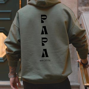 Könnte beinhalten: Olivgrüner Kapuzenpullover mit dem Wort "PAPA" in Schwarz vertikal auf dem Rücken gedruckt. Unter dem Wort "PAPA" steht der Text "est.2026". Der Hoodie hat eine Kapuze und lange Ärmel.