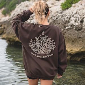 Könnte beinhalten: Ein braunes Sweatshirt mit einem weißen Lotusblumen-Design auf dem Rücken. Der Text "Yoga is cheaper than therapy" befindet sich unter der Blume. Das Sweatshirt wird von einer Person getragen, die sich in der Nähe von Wasser befindet.