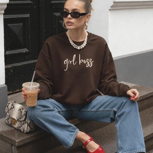 May include: A brown sweatshirt with the text "girl boss" in a cursive font. The person is wearing blue jeans, red shoes, sunglasses, and a pearl necklace. A snakeskin print purse and a drink with a straw are also visible.