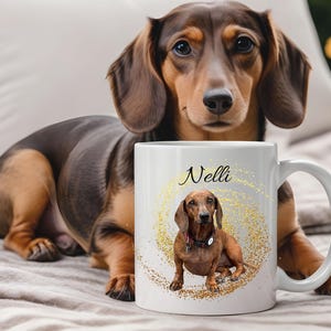 Könnte beinhalten: Eine weiße Keramiktasse mit einer Dackel-Illustration und dem Namen "Nelli" in Schreibschrift. Die Tasse hat ein goldenes Glitzerdesign um das Hunde-Bild. Im Hintergrund ist ein echter Dackel zu sehen.