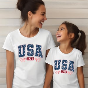 Könnte beinhalten: Weiße T-Shirts mit dem Wort "USA" in blauen, sternförmigen Buchstaben und der Zahl "1776" in Rot. Eine Frau und ein Kind tragen die T-Shirts und lächeln sich an.