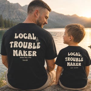 Könnte beinhalten: Zwei schwarze T-Shirts mit dem cremefarbenen Schriftzug LOCAL TROUBLE MAKER in einer Retro-Schriftart und einem Symbol für gekreuzte Finger. Das größere Shirt trägt den Text Since Day One DAVID, das kleinere Shirt zeigt MAXI est.2022.