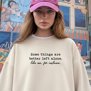 Peut inclure: Une personne portant un sweat-shirt beige clair avec le texte "Some things are better left alone. like me, for instance." et une casquette de baseball rose avec du texte blanc. L'arrière-plan présente une fresque colorée.