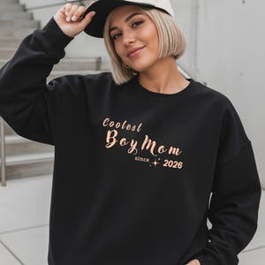Könnte beinhalten: Ein schwarzes Sweatshirt mit Rundhalsausschnitt und dem roségoldenen Schriftzug Coolest Boy Mom since 2026 sowie kleinen Sternakzenten. Das Kleidungsstück hat lange Ärmel und eine lockere Passform. Eine zweifarbige Baseballkappe mit weißer Krone ist zu sehen.