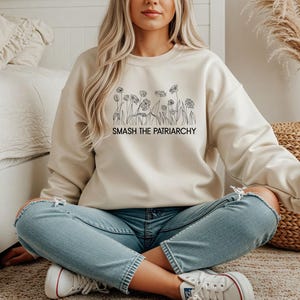 Könnte beinhalten: Cremefarbenes Sweatshirt mit schwarzem Blumendesign und dem Text "SMASH THE PATRIARCHY." Das langärmlige Oberteil wird mit hellblauen Jeans und weißen Turnschuhen kombiniert. Die Person sitzt auf einem Teppichboden.