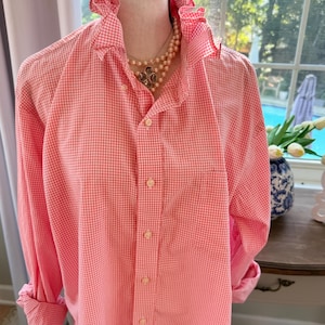 May include: A long-sleeved button-down shirt featuring a coral and white gingham pattern. The shirt has a ruffled collar, a front button placket with light-colored buttons, and a single chest pocket on the left side.