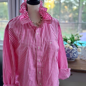 May include: A pink and white gingham button-down shirt with a ruffled collar and cuffs. The long-sleeved shirt is displayed on a mannequin. A pearl necklace is visible. The shirt has a pocket on the left side.