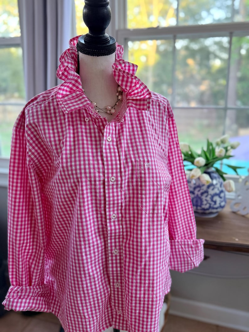 May include: A pink and white gingham button-down shirt with a ruffled collar and cuffs. The long-sleeved shirt is displayed on a mannequin. A pearl necklace is visible. The shirt has a pocket on the left side.