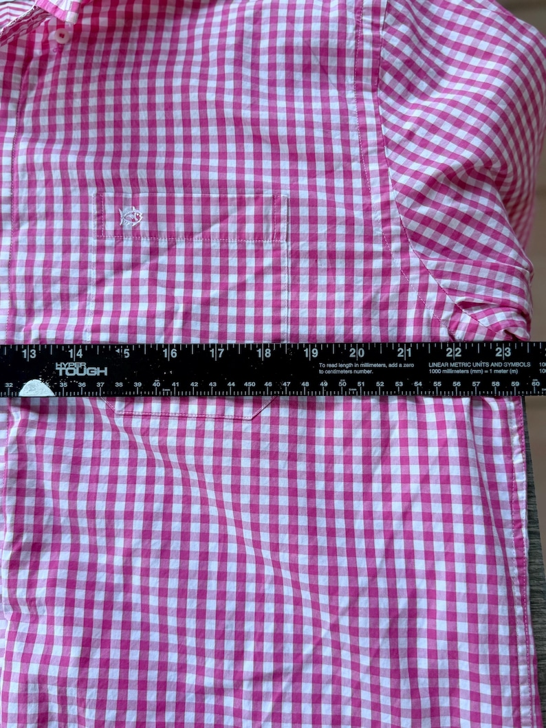 May include: A pink and white gingham-patterned shirt with a button-down collar and a chest pocket. The shirt has a small embroidered logo on the pocket. The ruler shows the shirt is approximately 20 inches wide.