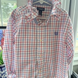 May include: A long-sleeve button-down shirt featuring a white base with a red and navy blue plaid pattern. The shirt includes a ruffled collar, a front button placket, and a small blue embroidered logo on the chest. The label reads CHAPS size M 10-12.