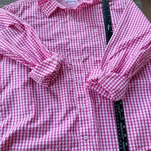 May include: A pink and white gingham button-down shirt with a ruffled collar and cuffs. The long-sleeved shirt has a small pocket on the chest and a classic collar. The shirt is made of a lightweight fabric.