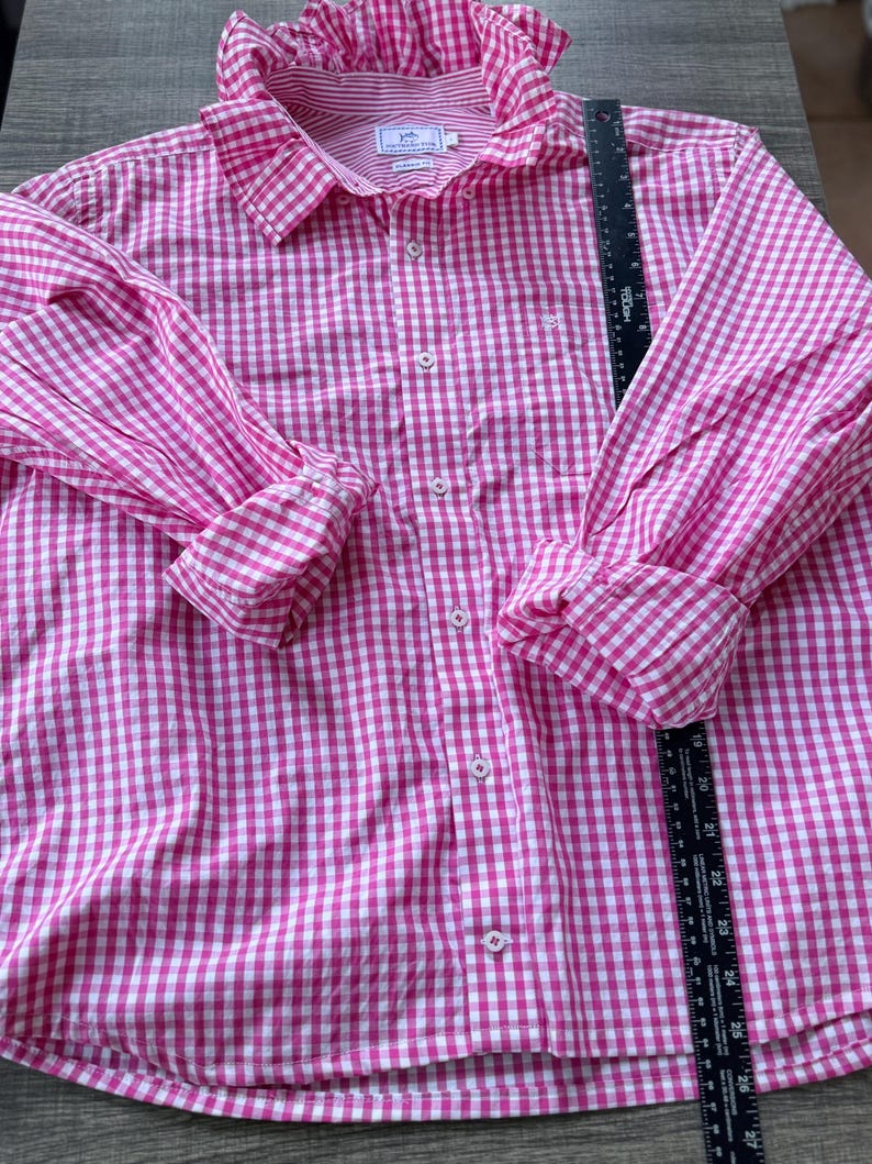 May include: A pink and white gingham button-down shirt with a ruffled collar and cuffs. The long-sleeved shirt has a small pocket on the chest and a classic collar. The shirt is made of a lightweight fabric.
