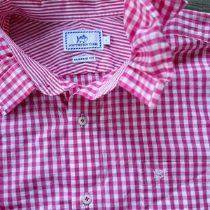May include: A pink and white gingham-patterned shirt with a ruffled collar. The shirt features a button-down front, a chest pocket with a small embroidered fish, and a label that reads "SOUTHERN TIDE CLASSIC FIT".